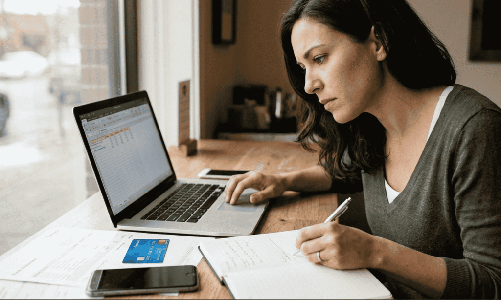 Woman tracking finances on a laptop and notebook at a cafe table.